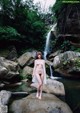 A naked woman standing on a rock in front of a waterfall.
